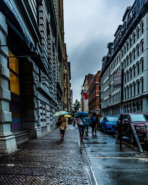 Urban street scene with pedestrians using umbrellas in rainy weather, flanked by tall buildings.