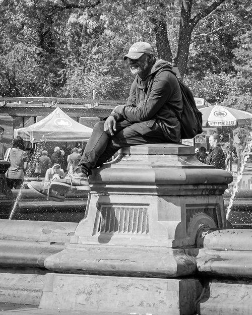 Man sitting on a fountain ledge in a park, wearing casual clothes and a cap, with people and trees in the background.