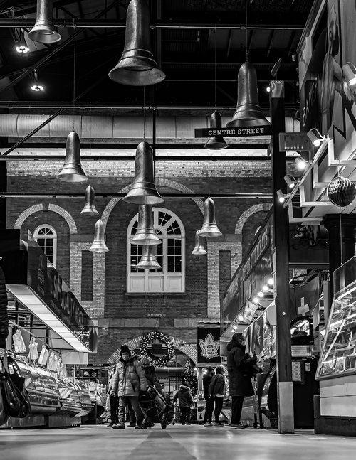 Black and white photo of an indoor market scene with shoppers walking between vendor stalls. The ceiling features large, bell-shaped lights. A brick wall with arched windows in the background indicates an urban setting. The market is decorated with h