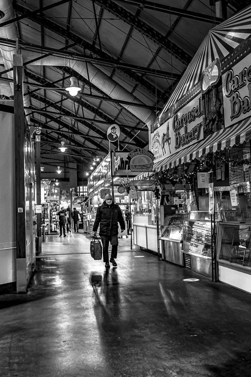 Black and white image of an indoor market with a high ceiling, featuring stalls and a person walking with a bag. "Carousel Bakery" is visible on the right.