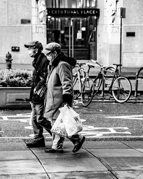 Two people wearing masks walking on a city street near a row of bicycles, carrying shopping bags, with a building labeled 'Cathedral Place' in the background.