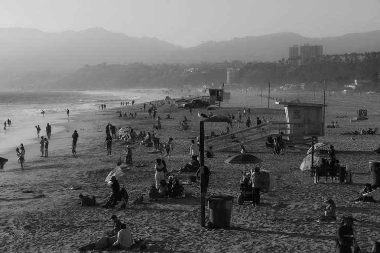 Crowded beach scene in black and white with people relaxing on the sand, walking along the shoreline, and a lifeguard tower in the background. Distant hills and buildings are visible.
