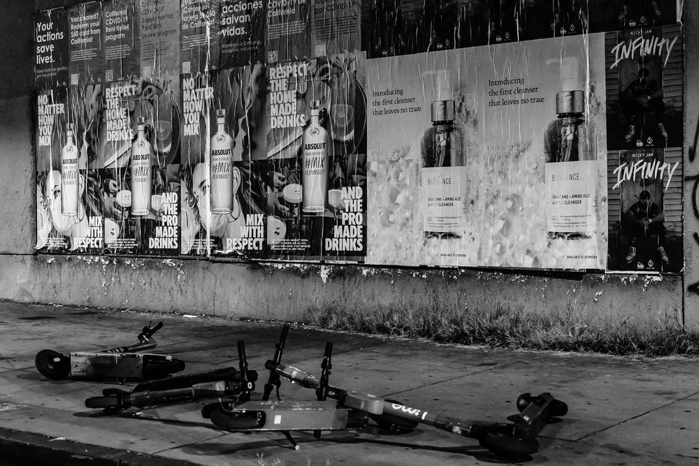 Black and white image of various posters on a wall with fallen scooters on the sidewalk below. The posters include advertisements for vodka, a skin cleanser, and a musical release. The ground is slightly wet and uneven.