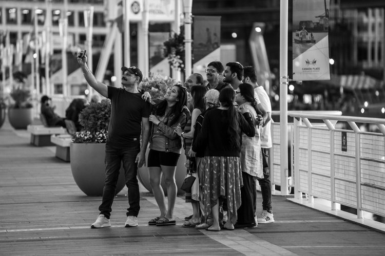 Group of people taking a selfie on a crowded urban outdoor walkway, with city lights and banners in the background, in black and white.
