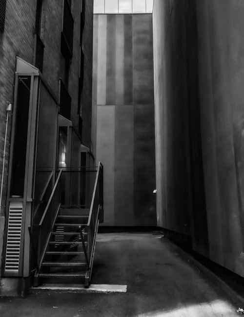 Black and white photo of an industrial alley with metal fire escape stairs and tall building walls.