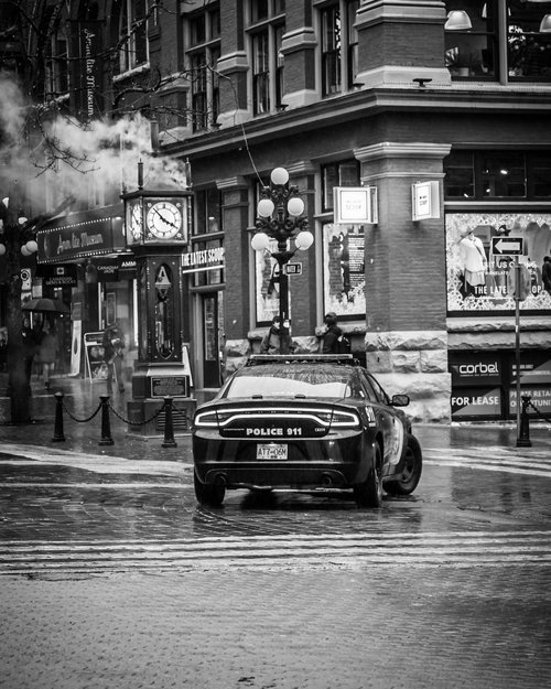 Black and white photo of a police car on a wet city street, near a vintage clock and storefronts in a historic district.