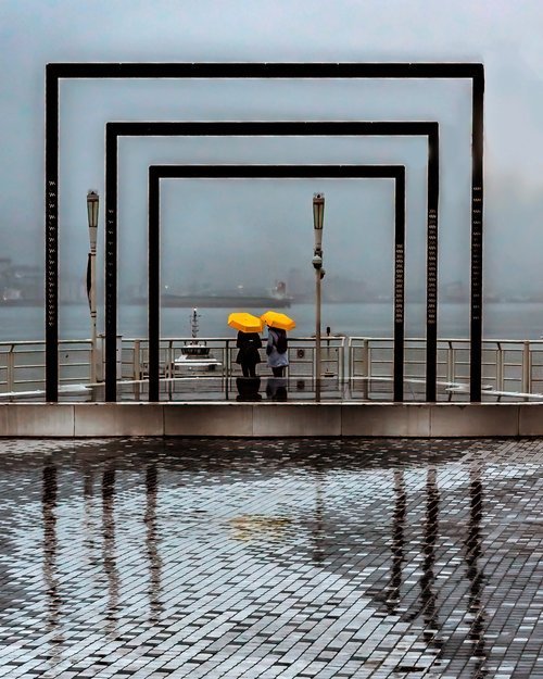 Two people holding yellow umbrellas stand in the rain under large black rectangular frames by a waterfront, with a brick-paved walkway reflecting the scene.