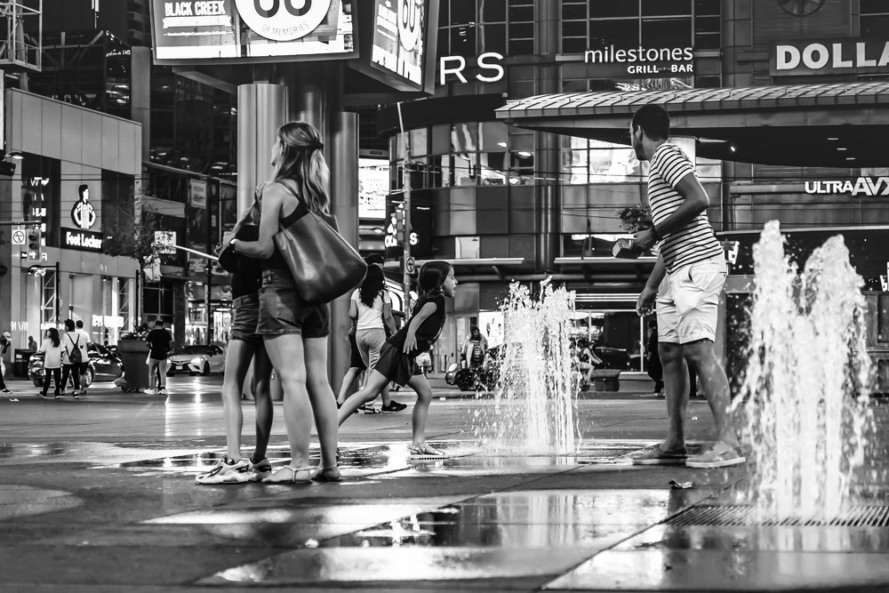 Black and white photo of people in a city square with water fountains, surrounded by commercial buildings and advertisements. Several individuals are interacting with the fountains.