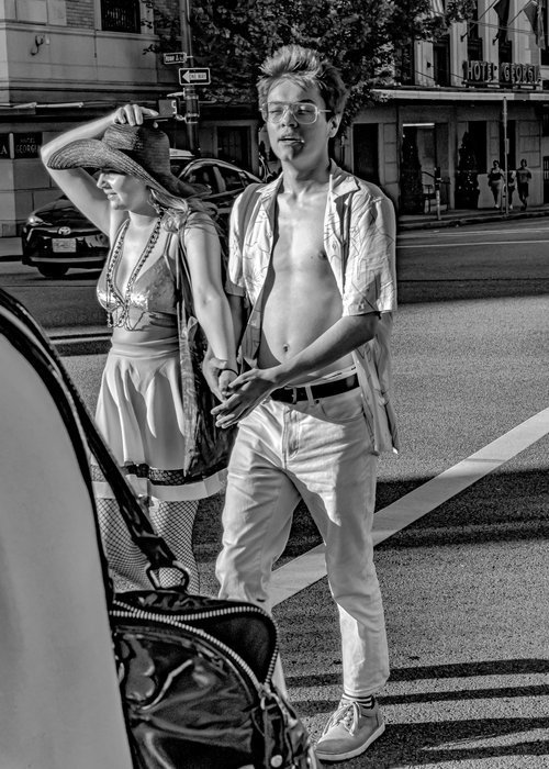 Two people walking across a street, one wearing an open shirt and glasses, and the other in a wide-brimmed hat and dress. It's a black and white photo, with a vintage look.