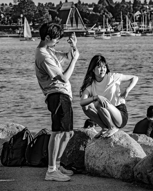 A black and white photo of a young man taking a picture of a woman crouching on a rock by the water, with sailboats and trees in the background.