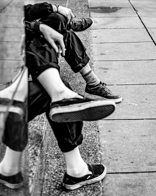 Black and white photo of two people sitting on a curb, wearing casual shoes and rolled-up pants. One person wears white socks and another wears patterned socks. Partial reflections are visible on a glass surface.