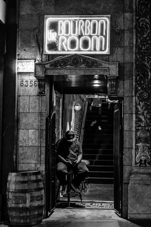 Black and white photo of a dimly lit entrance to "The Bourbon Room" with a neon sign above. A person is sitting on a stool in the doorway, and stairs leading upward are visible inside. A wooden barrel is placed outside, contributing to the rustic dec
