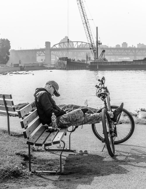 A person sitting on a bench near a river, with feet propped up on a bicycle. A bridge and construction crane are visible in the background.