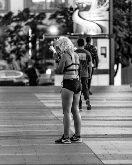 Black and white photo of a person standing on a sidewalk in a city, reading a paper. They are wearing shorts and a top. Other pedestrians are visible in the background walking away.