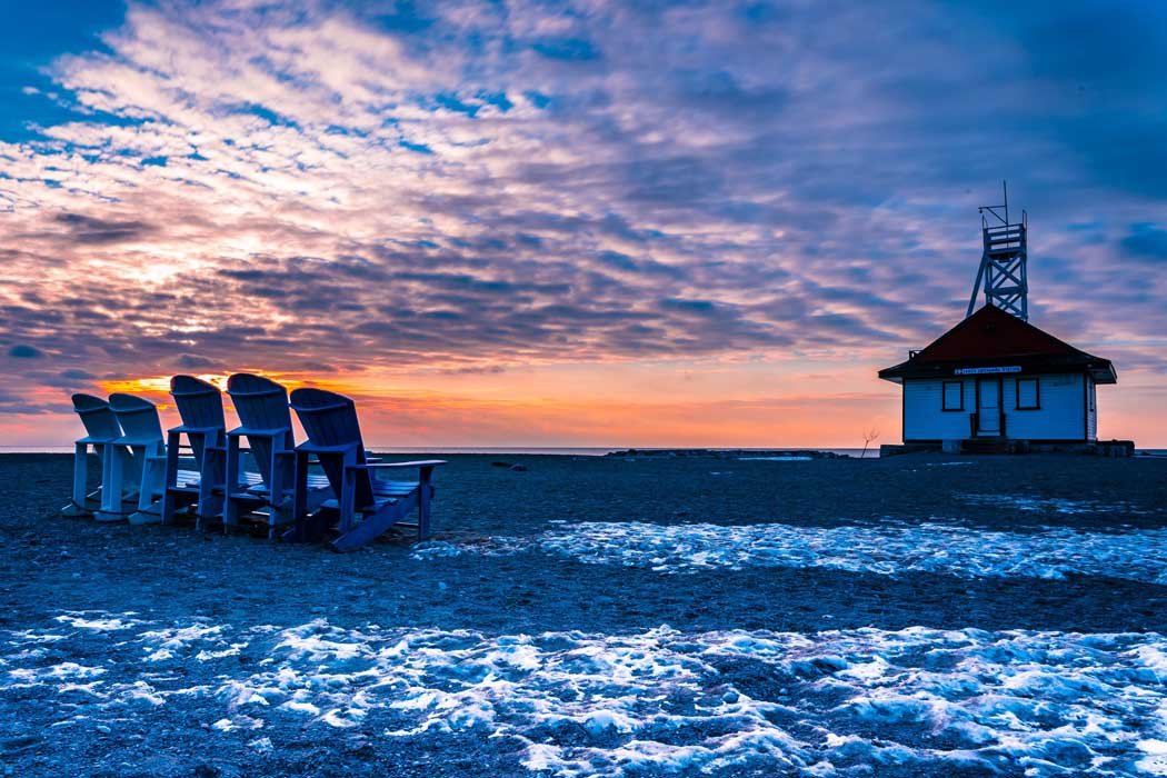 Beach sunrise with Adirondack chairs and a lifeguard station, snowy foreground.