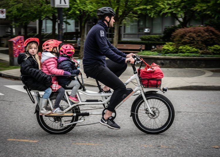 Man cycling on a cargo bike with three children in helmets riding on the back seat. A red bag is attached to the bike. Urban setting with trees and buildings in the background.