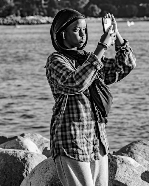 Person in a plaid shirt taking a selfie near a body of water with a mobile phone.