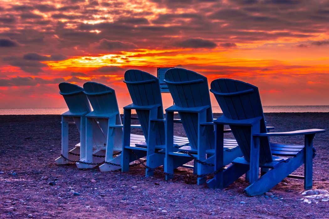 Five Adirondack chairs on a beach facing a dramatic sunset with orange and purple skies.