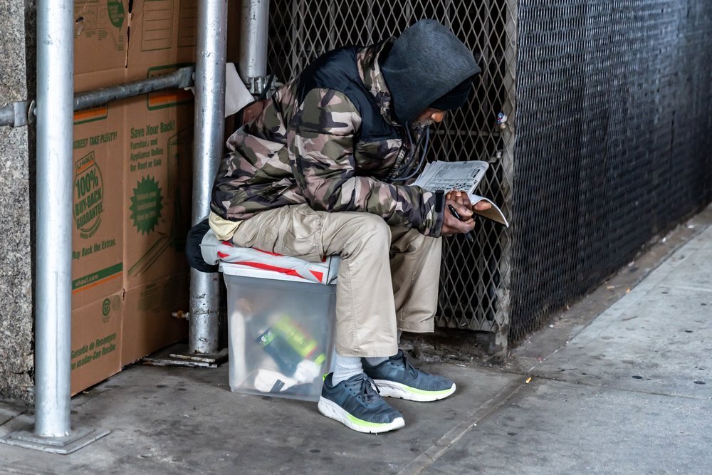 A person in a camouflage jacket and hood sits on a plastic container on a sidewalk, under scaffolding next to cardboard boxes. They are reading or writing in a booklet.
