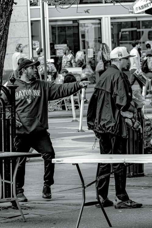 Two men in a street scene, one reaching into the other's pocket, with people and a store entrance in the background.