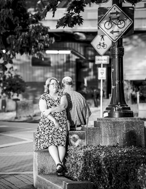 A woman sitting on a stone ledge next to a man, under a bicycle route sign in an urban setting. The scene is in black and white, capturing a relaxed moment.
