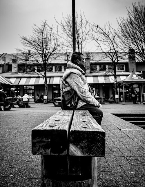 Black and white photo of a person sitting on a wooden bench in an outdoor plaza, with buildings and bare trees in the background.