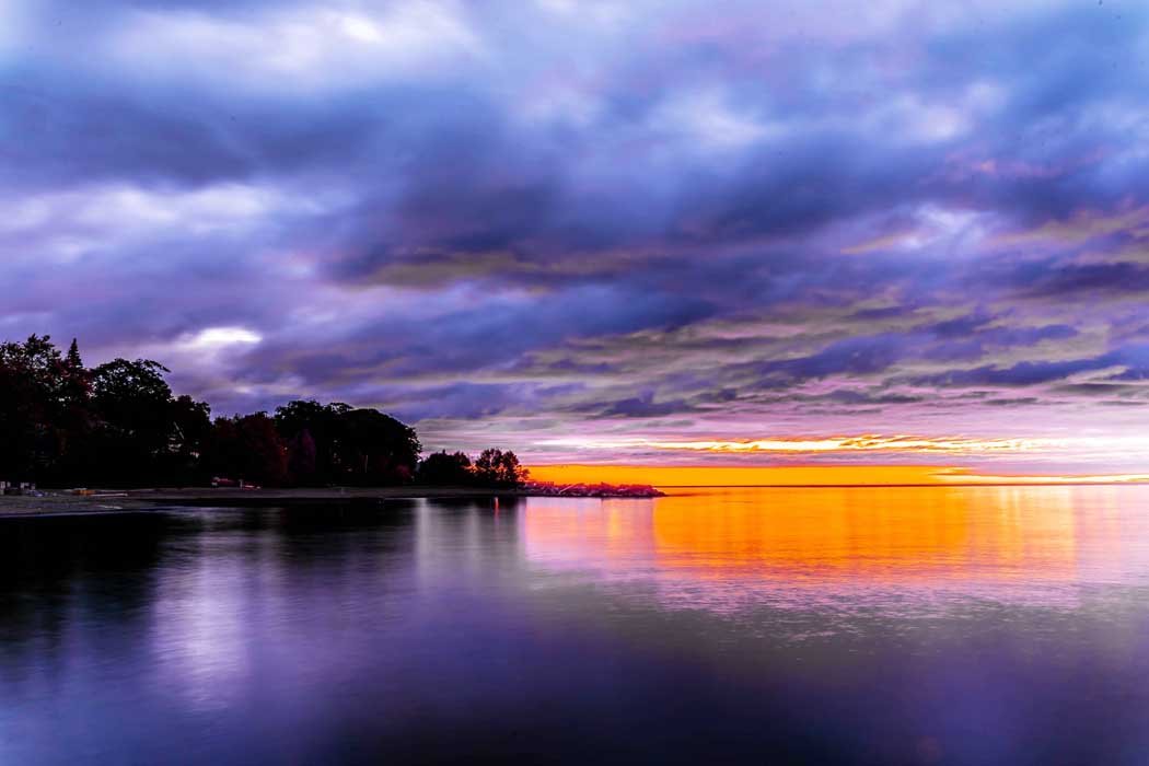Sunset over a lake with vibrant purple and orange sky, silhouetted trees on the left, and calm water reflecting the colors.