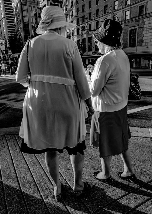 Two elderly women wearing hats, walking on a city street. They are dressed in light jackets and skirts, standing on a crosswalk. The scene is in black and white.