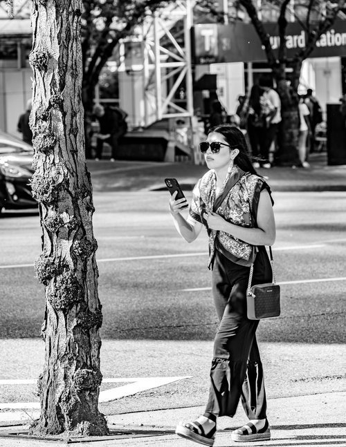 Black and white image of a woman walking on a street, holding a smartphone. She is wearing sunglasses, a patterned scarf, and carrying a small handbag. There is a tree in the foreground and several people and signs in the background.