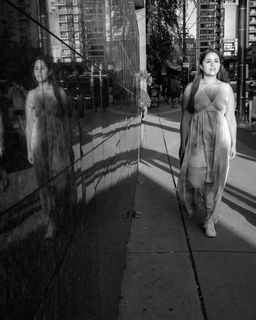 Woman walking on city sidewalk with reflection in glass wall in black and white photo.