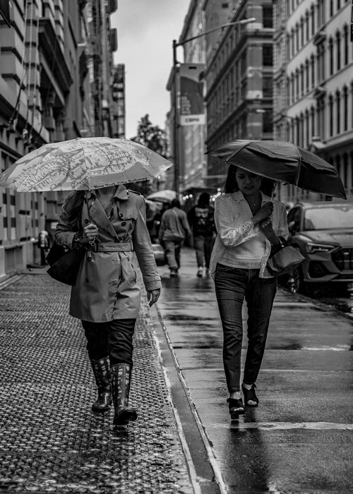Black and white photo of two people walking on a rainy city street with umbrellas.