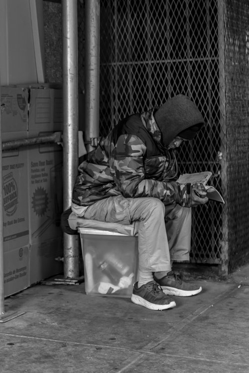 Black and white photo of a person wearing a hooded jacket sitting on a plastic container, reading a newspaper on a city sidewalk.