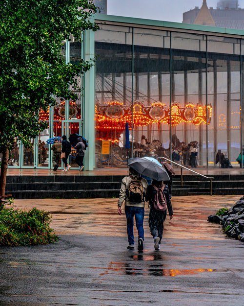A couple walking under an umbrella on a rainy day towards a glass building housing a brightly lit carousel.