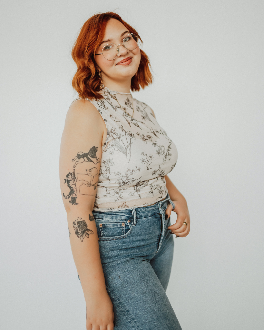 A person with long red hair and glasses stands smiling in a hair salon, wearing a black apron labeled "Copper Lane hair studio." Behind them, shelves with hair products and a potted plant are visible.