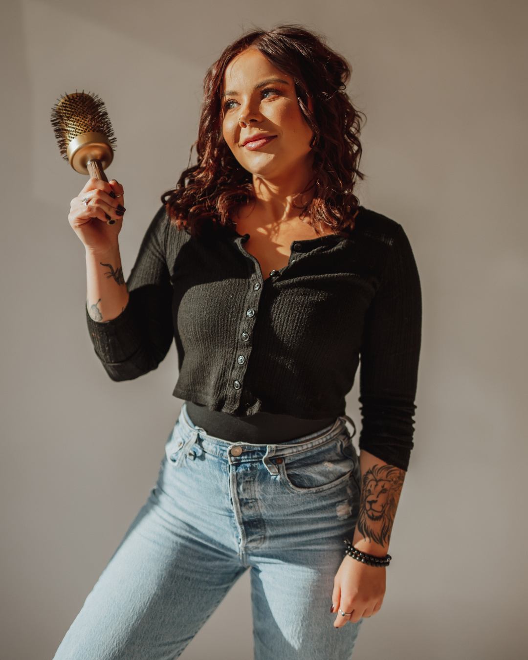 A young woman with long wavy red hair sitting on a salon chair inside a hair salon, smiling at the camera, wearing a black top, a sleeveless denim jacket, black pants, with tattoos on her left arm, in front of a mirror with salon shelves and chairs visible in the background.