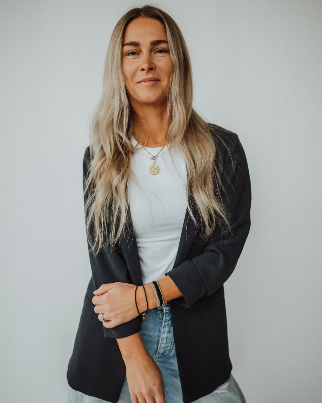 Person with long blonde hair wearing a black turtleneck, standing near a chair in a modern indoor setting with mirrors.