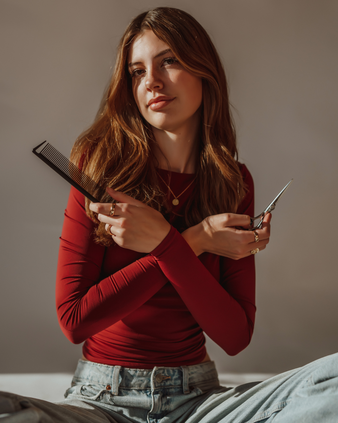 A young woman with curly brown hair sitting on a black salon chair in a hair salon, wearing a black sleeveless top, light blue jeans, a pearl necklace, and matching pearl bracelets. She is smiling and looking at the camera, with a bookshelf and white wall in the background.