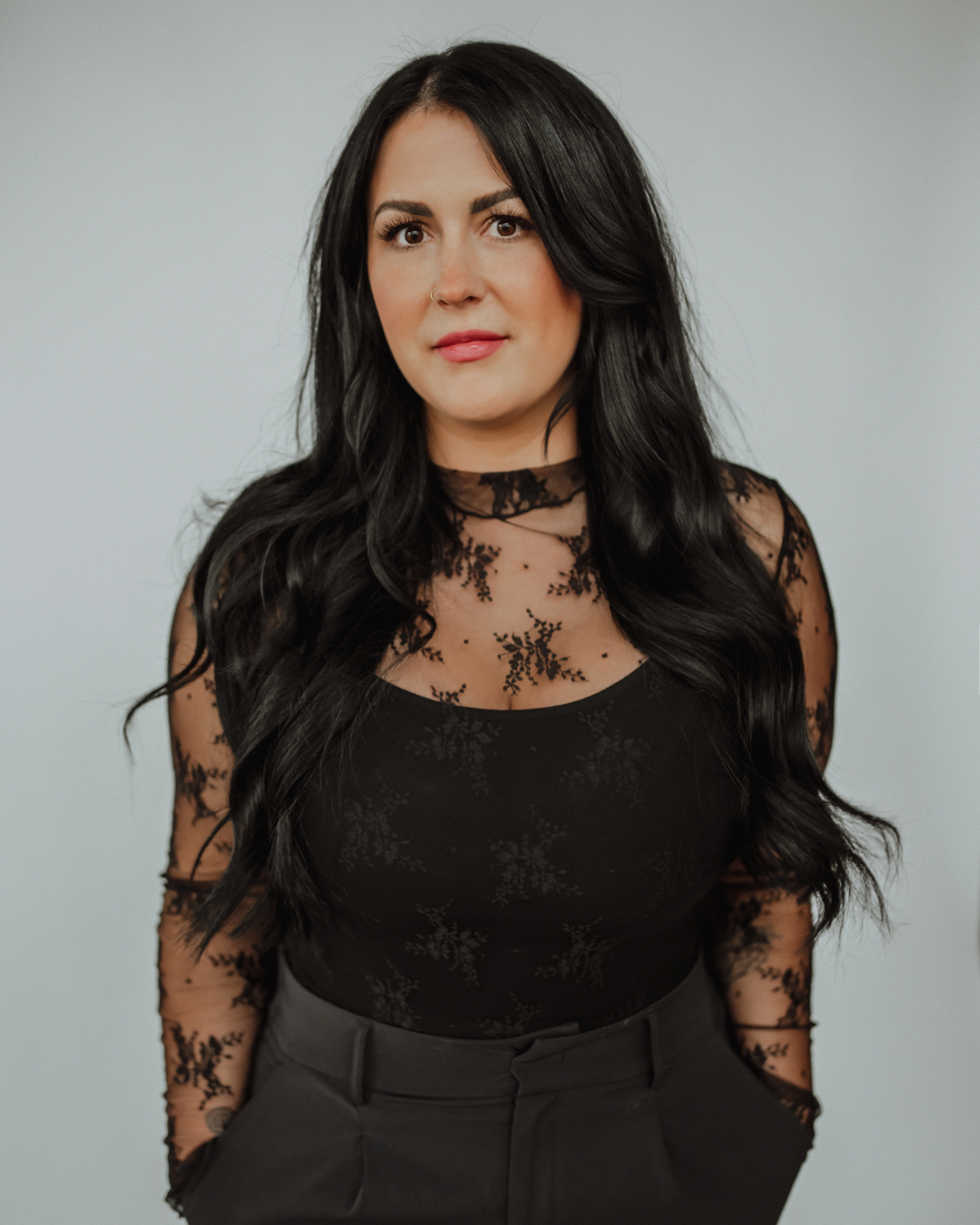 Woman with long black hair in a salon, wearing black top and jeans, sitting on a chair while another person cuts hair in the background.