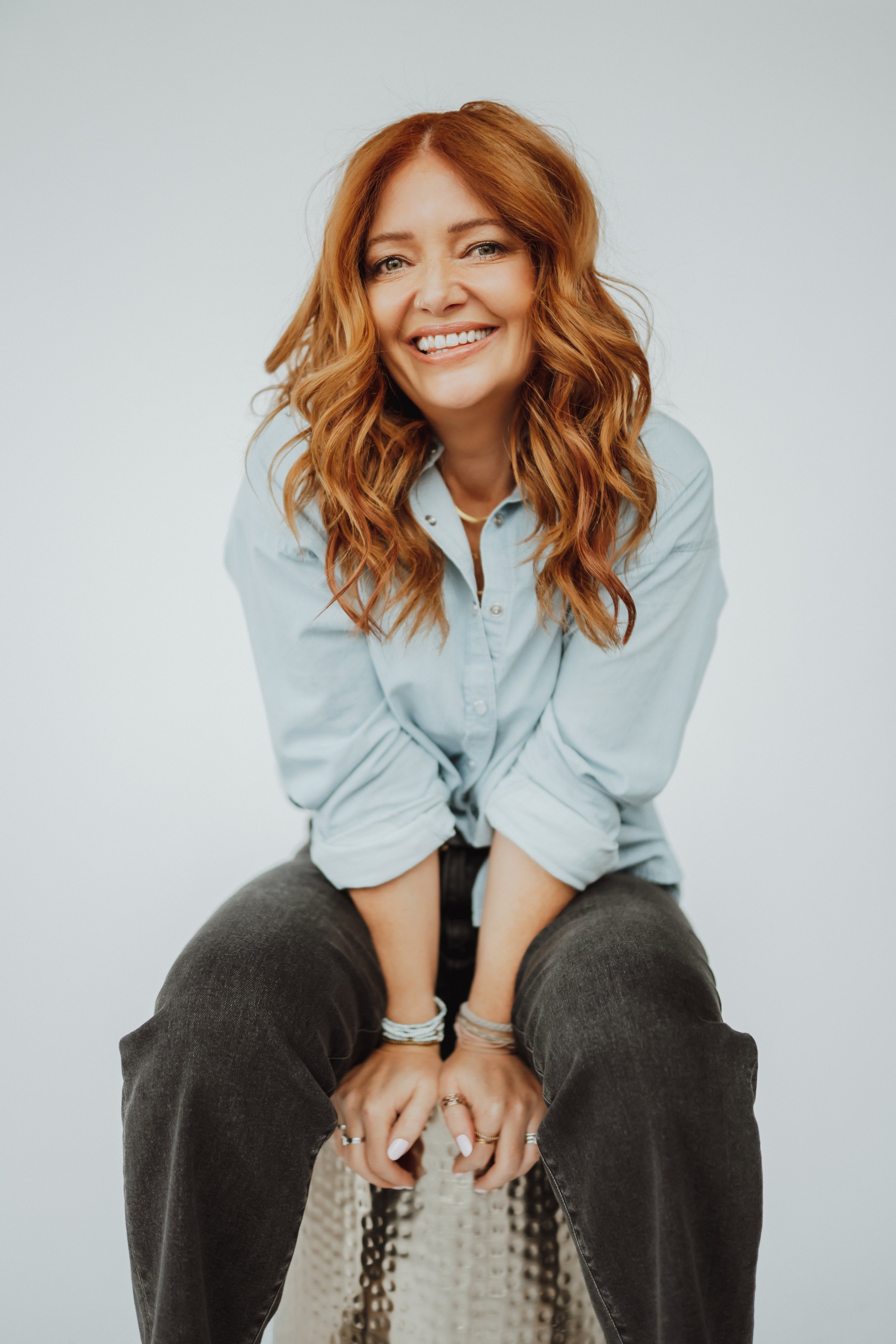 Woman with wavy brown hair wearing a beige sweater and blue jeans in a clothing store, smiling in front of mirrors and racks of clothes.