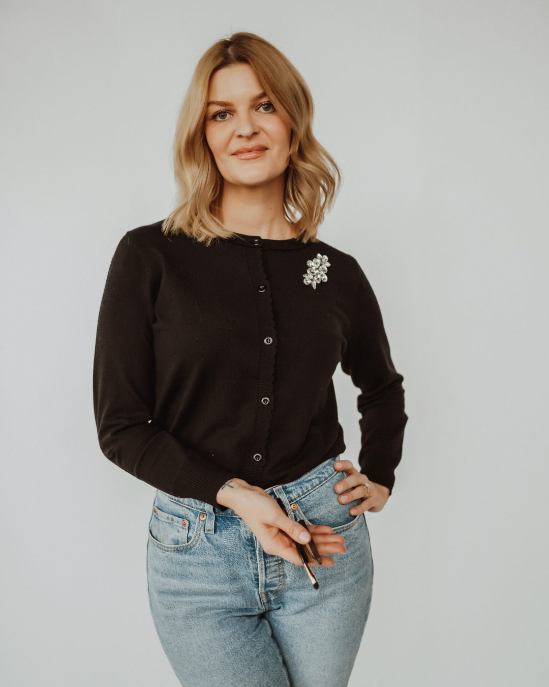 A woman with long wavy hair wearing a white blouse and layered necklaces smiles inside a modern interior setting.