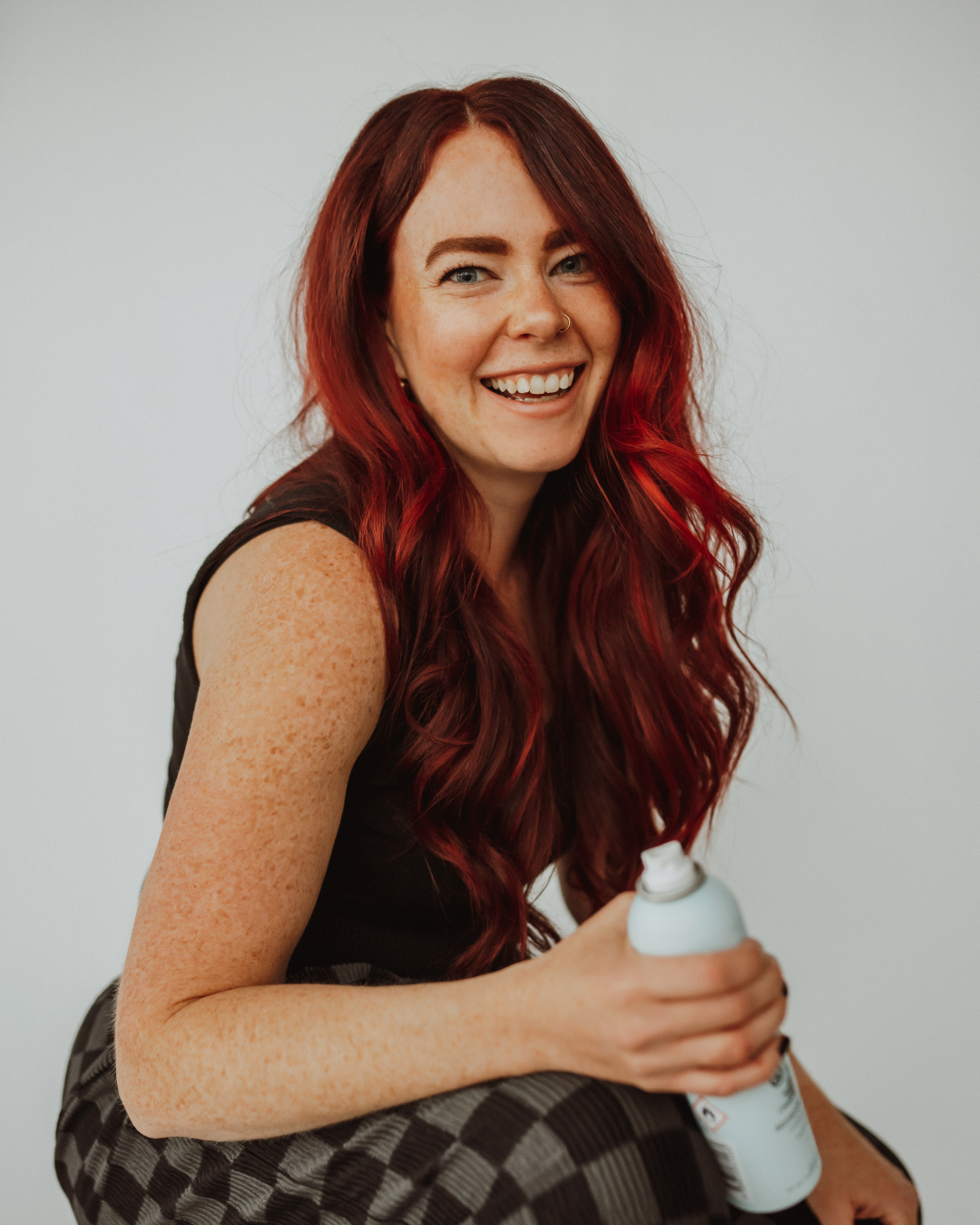 Person with long red hair smiling, wearing black clothing, standing indoors.