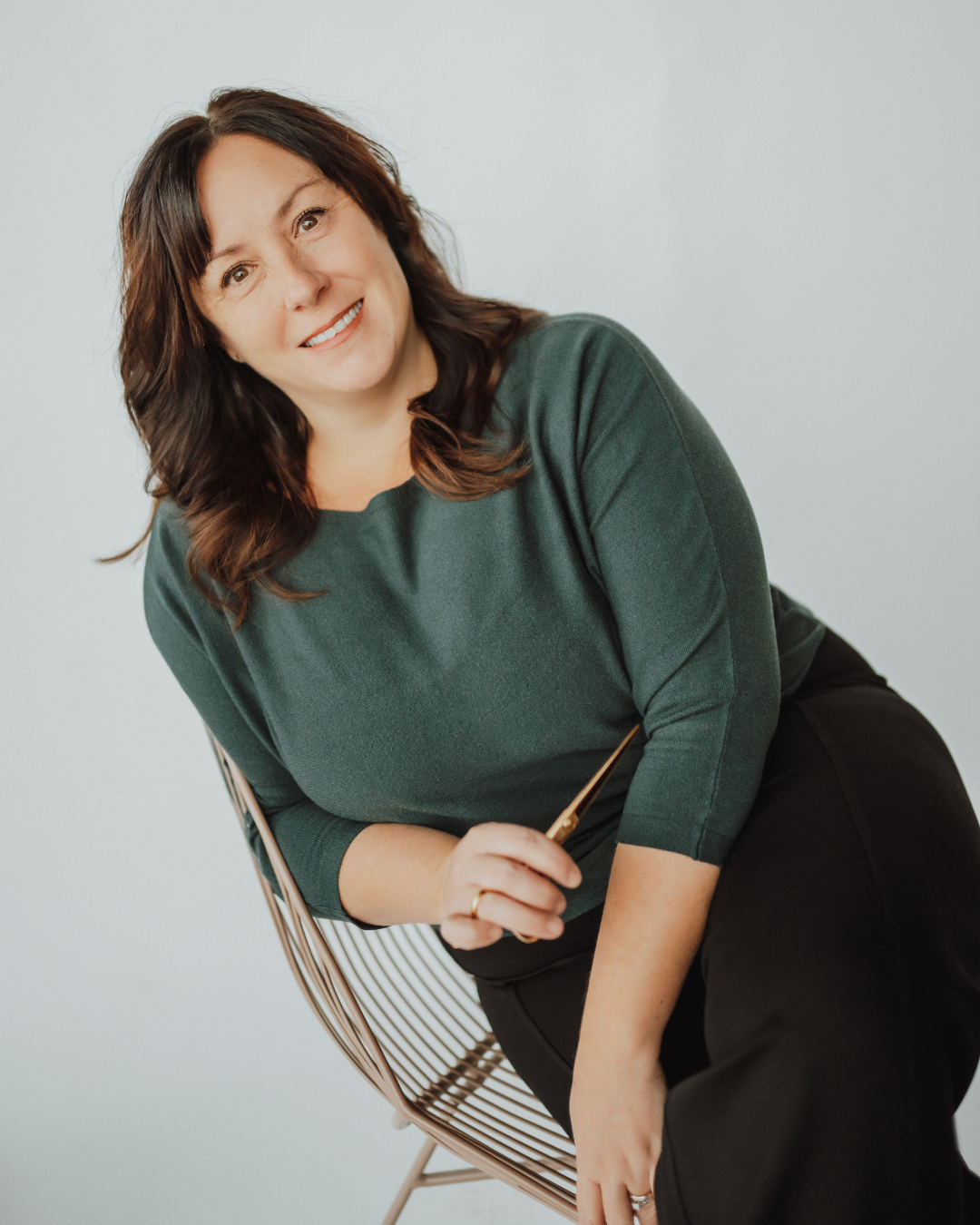 Woman sitting in a salon chair with styled hair, wearing a black top, smiling, and a shelf with hair products in the background.