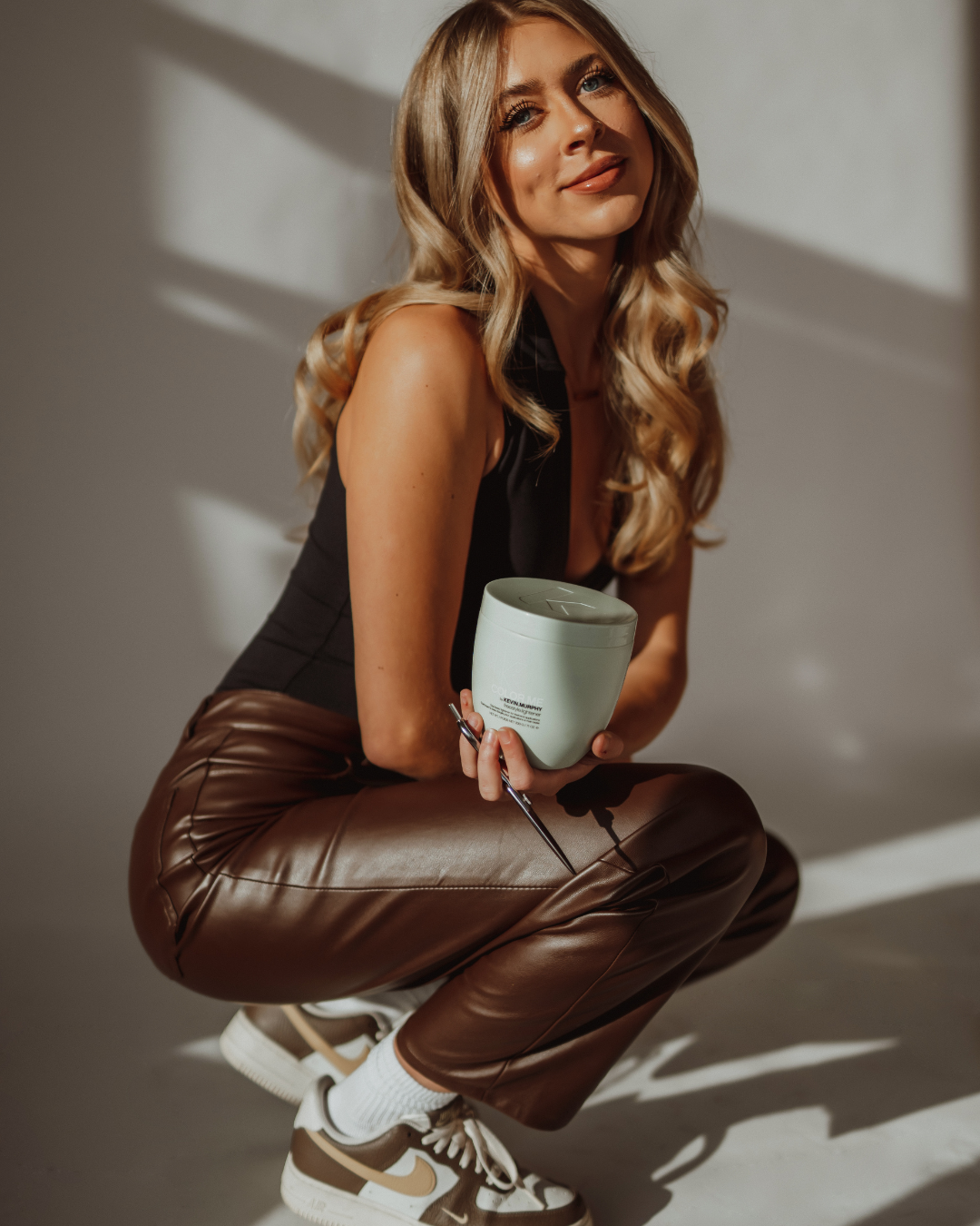 Woman holding an orange Amika branded bottle, standing in a hair salon with a clock on the wall. An orange bag with a floral pattern is in the foreground.