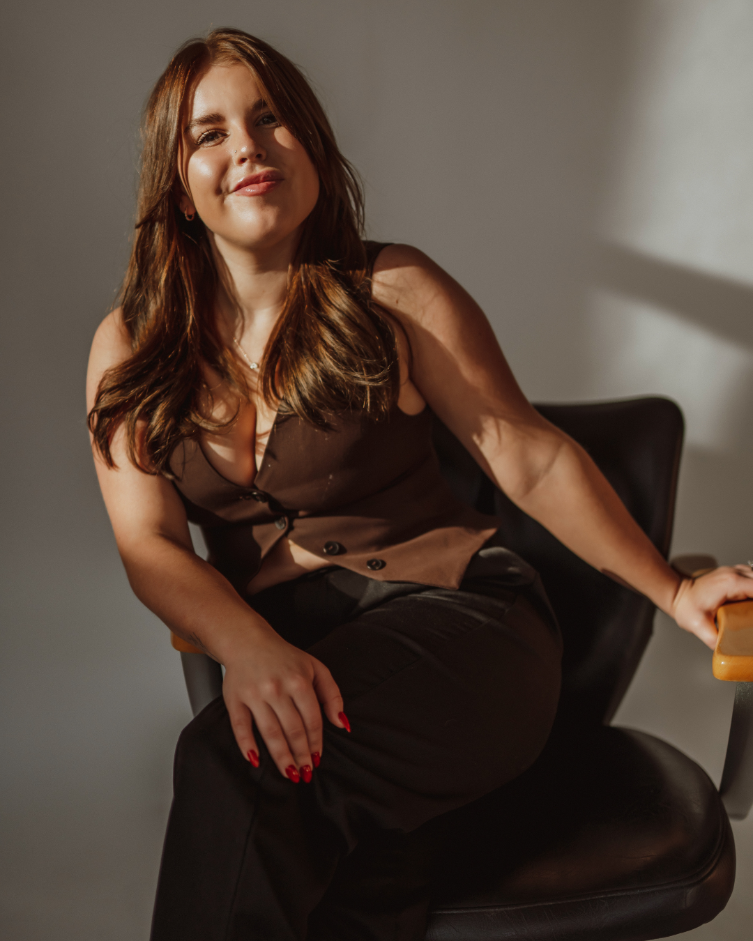 Woman in beige top and jeans standing in a salon, smiling, with shelves of hair products in the background and a reflection in the mirror.