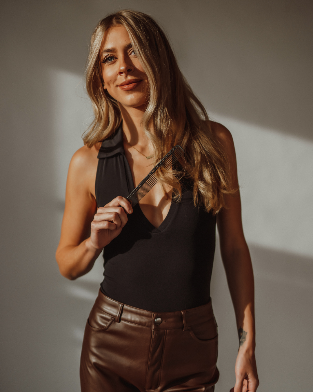 A woman with long wavy blonde hair sitting on a black salon chair inside a hair salon. She is smiling, wearing a brown long-sleeve top and light blue jeans.