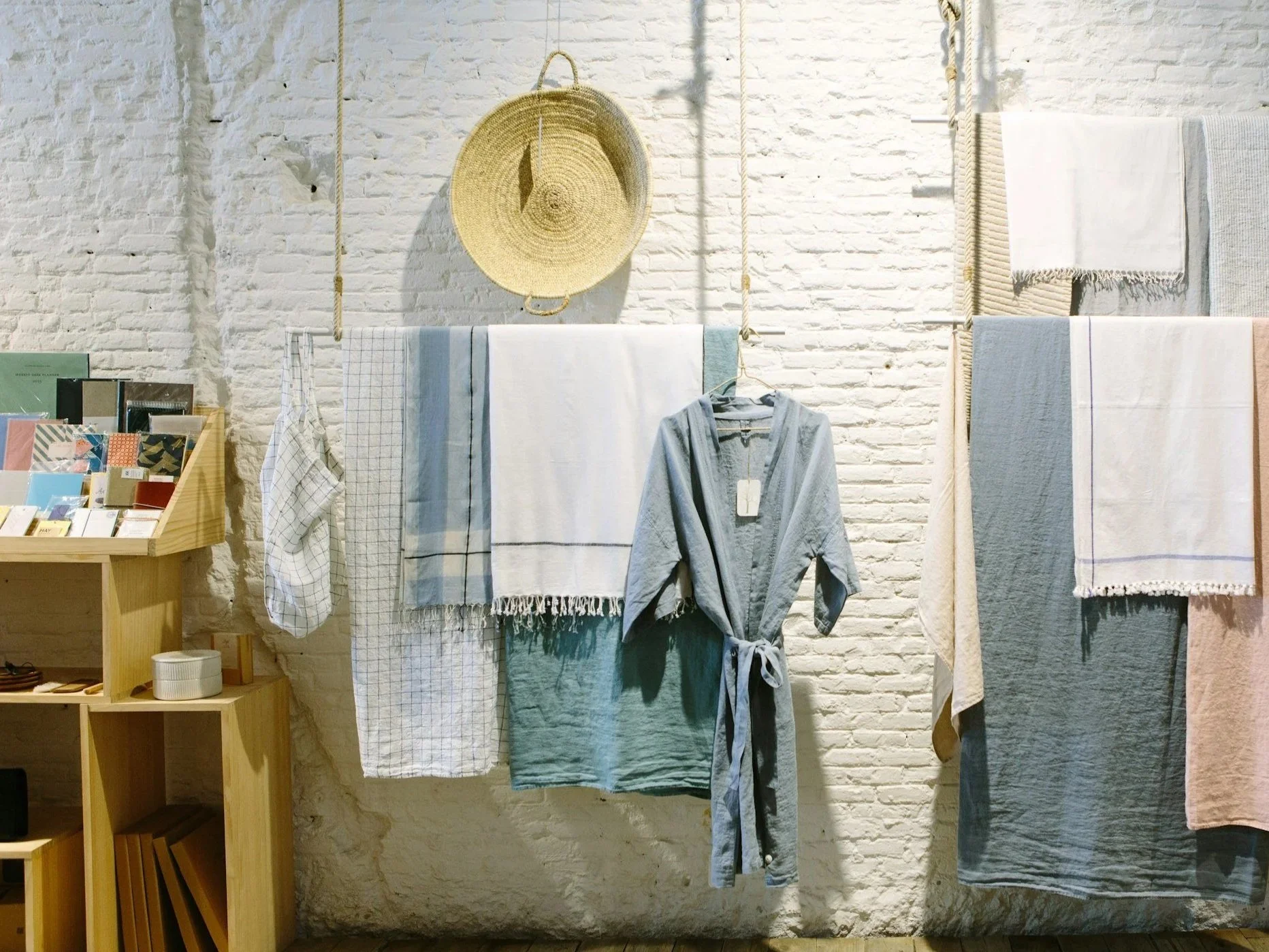 Présentation de textiles suspendus, notamment des serviettes, des chiffons et un sweat à capuche, avec un panier tissé sur un mur de briques blanches dans un magasin de détail ou d'articles pour la maison.