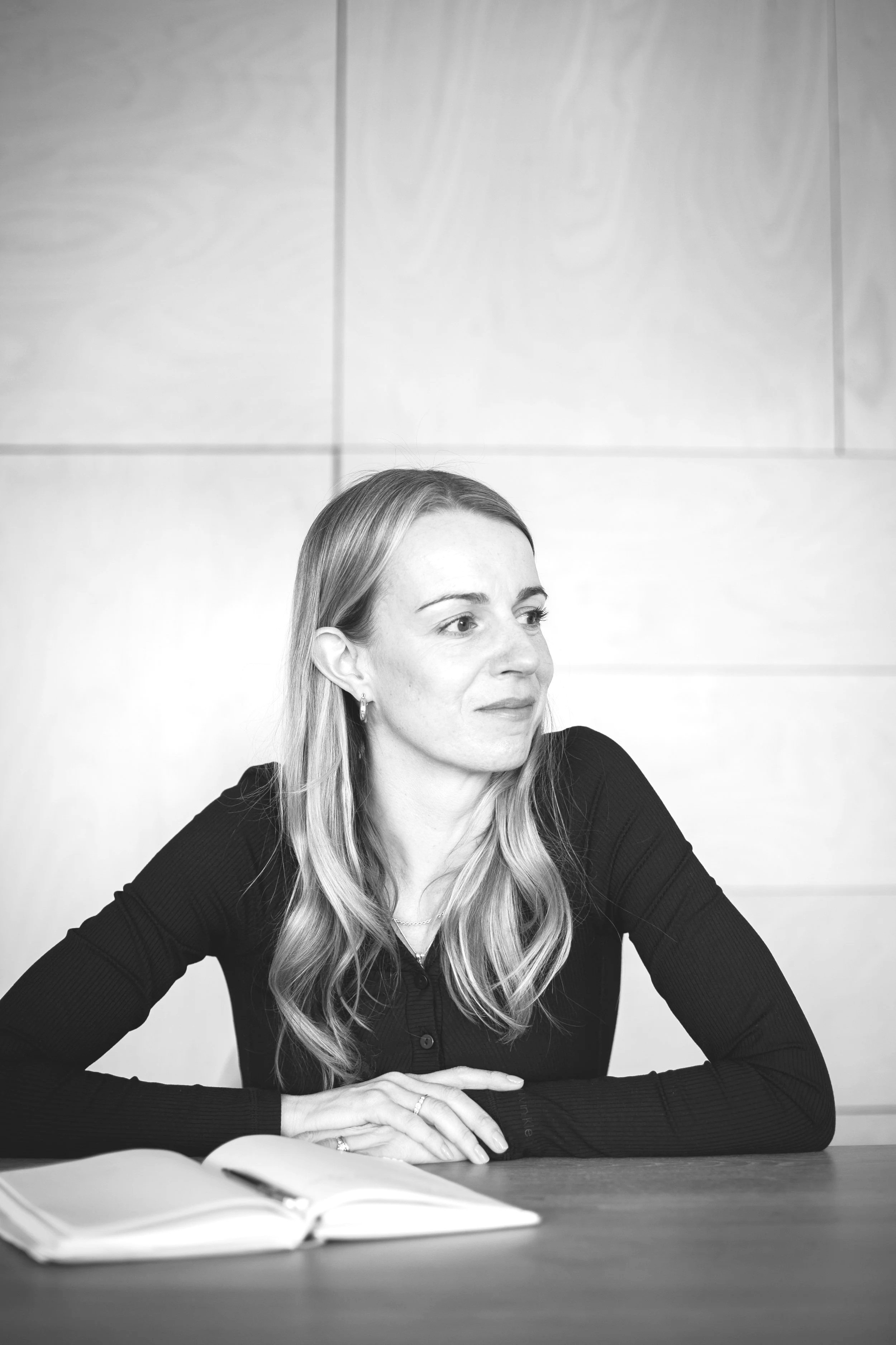 Image of Jo Matthews wearing earrings and a black top, sitting at a table with her arms crossed, looking to her left. An open book is in front of her on the table. The background contains light-coloured wall panels.