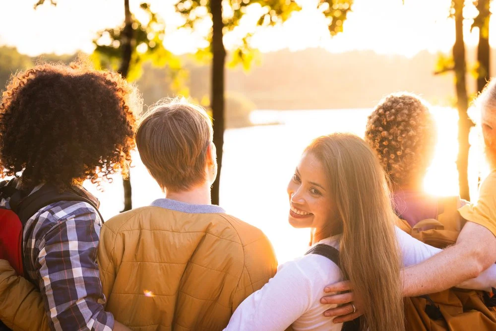 Dieses herzerwärmende Bild zeigt eine Gruppe von Freunden, die den Sonnenuntergang am See genießen. Mit dem Rücken zur Kamera stehen die Freunde in enger Umarmung und symbolisieren damit ihre starke Verbundenheit. Eine Frau dreht sich um