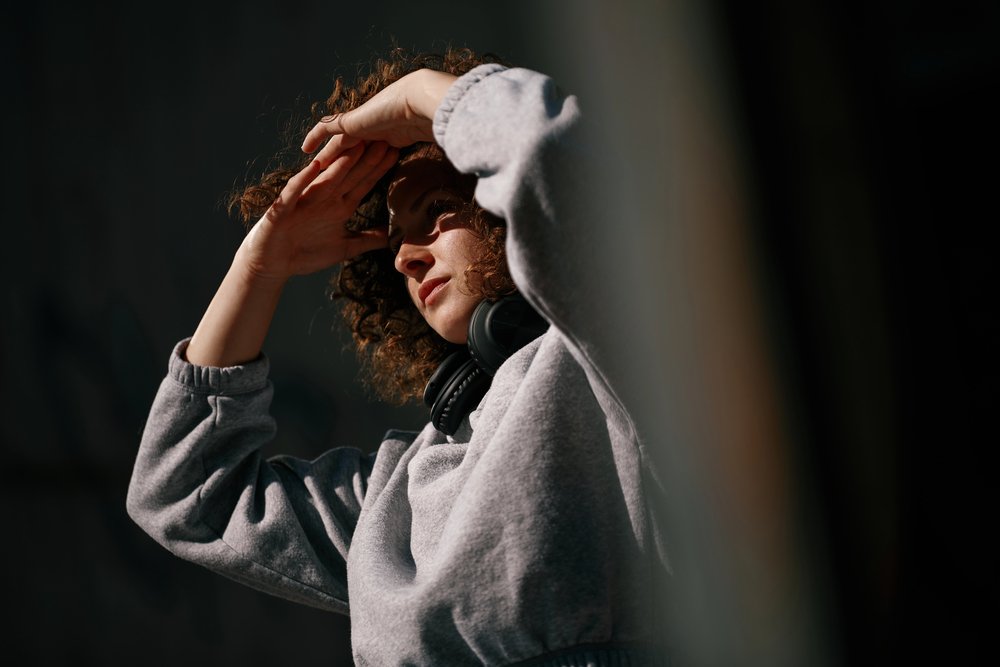 Eine junge Frau mit lockigem Haar und Kopfhörern um den Hals steht im Freien und blickt in Richtung Sonne.
