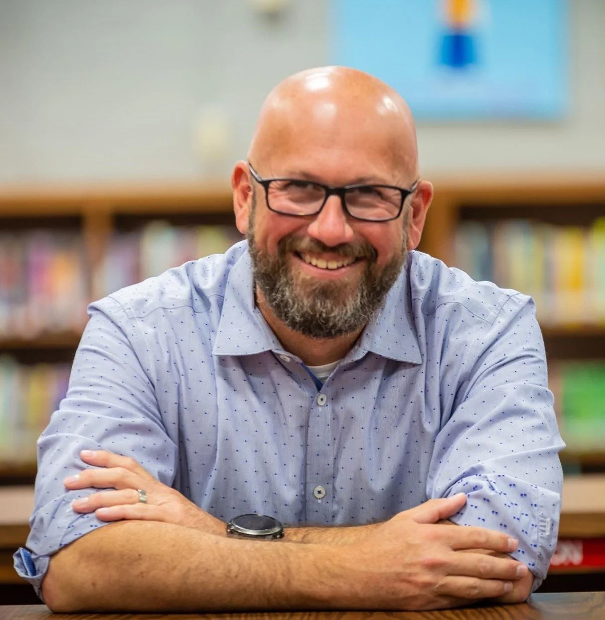 Mr. Reeves smiling, sitting at a table with arms crossed in a library.